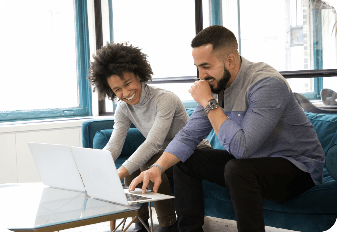 Two people sit on a blue sofa, smiling and working together on laptops at a glass coffee table in a bright office.